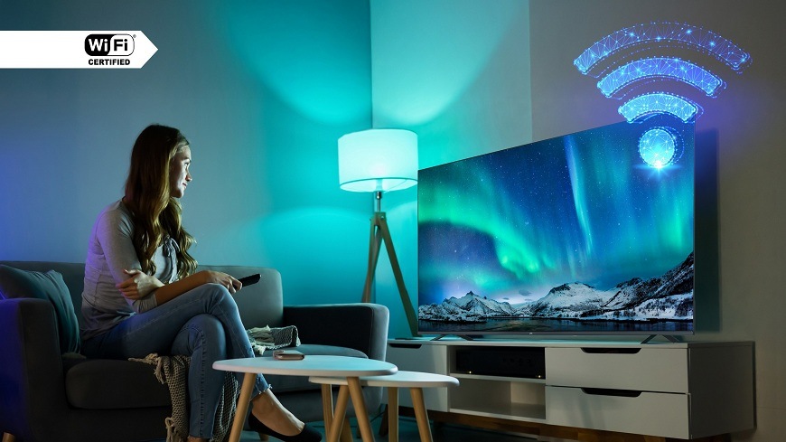 Televize JVC LT-65VDQ9435 Žena sedí na pohovce v modře nasvíceném obývacím pokoji, sleduje na Wi-Fi certifikované televizi JVC živý obraz polární záře, což zdůrazňuje bezdrátové připojení k internetu a kvalitu obrazu