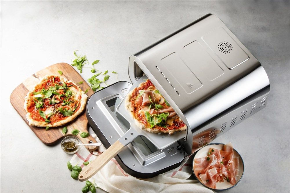 Pohled shora na pec na pizzu Domo GENIUS DO9289PZ, pec je otevřená, do pece je vložena lopatka, na níž leží pizza, v okolí pece leží ingredience a hotová pizza