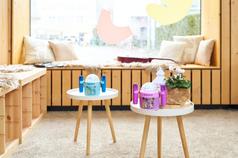 Dva dětské reproduktory GoGEN Bob a Bobek Disco s mikrofony, jeden v tyrkysové a druhý v růžové barvě, stojí na malých bílých stolcích v prosvětleném dřevěném interiéru. V pozadí je okenní parapet s polštáři a kytarou, vytvářející útulný koutek