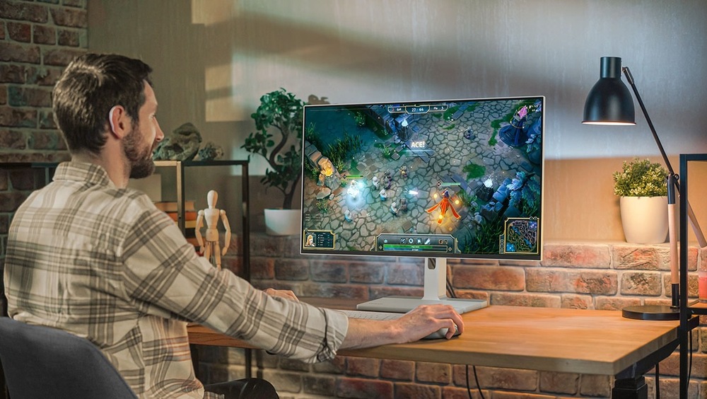 Monitor Lenovo L27h-4A šedý Muž v pruhované košili hraje videohru podobnou League of Legends na monitoru v útulné místnosti s cihlovými stěnami. Teplé osvětlení a rostliny vytvářejí uvolněnou atmosféru.