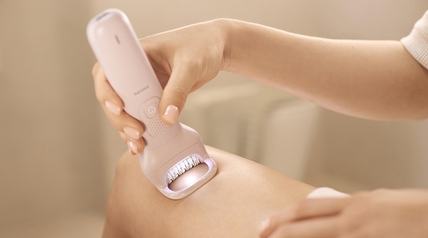 Epilátor Philips BRE718/00 v růžové barvě přiložený k pokožce nohy, detail epilační hlavice s osvětlením během odstraňování chloupků.