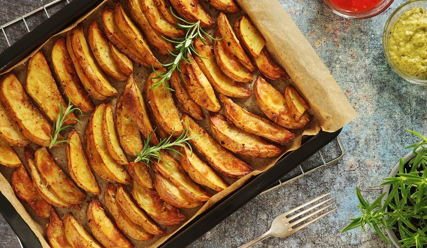 Pohled zblízka na plech plný zlatavých, křupavých pečených bramborových klínků s čerstvým rozmarýnem, připravených v troubě ETA 4788. Vedle plechu je vidět stříbrná vidlička, malé misky s kečupem a zelenou omáčkou a zelená bylinka v květináči