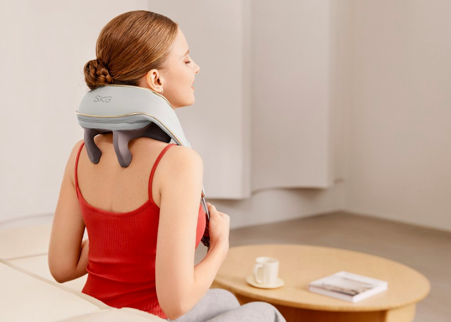 Masážní límec SKG H5 Mini šedý/bílý Žena používá masážní límec SKG H5 Mini na krk při relaxaci na pohovce