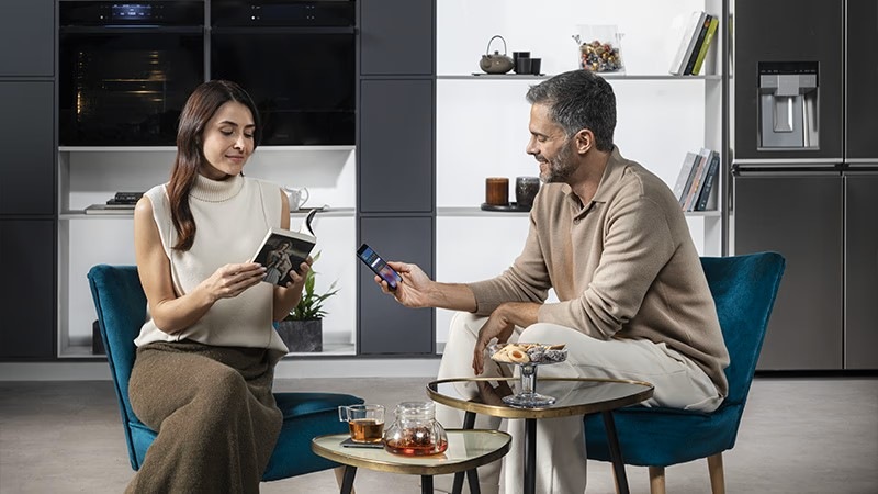 Haier H6 ID46G5SYTB dva lidé sedí u nízkého stolu a užívají si společné chvíle, zatímco ovládají troubu přes telefon