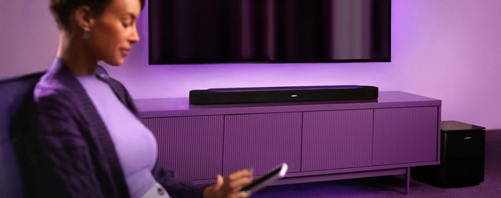 Soundbar Bose Lifestyle Ultra bílý Žena ovládá soundbar Bose Smart Ultra dálkovým ovladačem ve fialově nasvícené obývací pokoji.