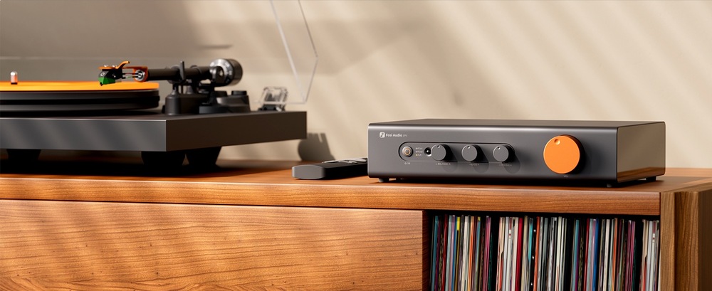 Zesilovač Fosi Audio ZP3 černý Předzesilovač Fosi Audio ZP3 s výrazným oranžovým ovladačem stojí na dřevěné skříňce vedle gramofonu a dálkového ovladače. Ve spodní polici jsou vinylové desky.