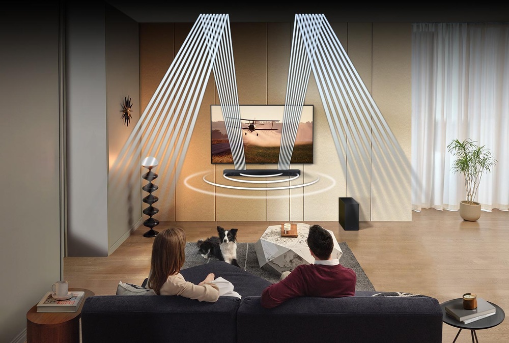 Soundbar Samsung HW-Q600C je umístněý na zdi pod televizí Samsung, před televizí sedí dva lidé na pohovce a sledují televizi, na koberečku před televizí leží pes, ze soundbaru vycházejí bílé čáry znázorňující zvuk