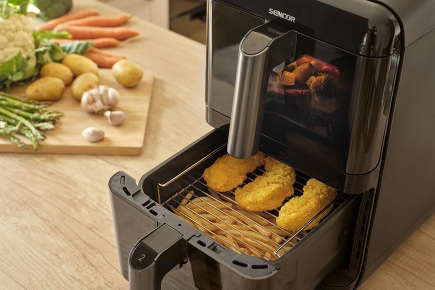 Fritéza horkovzdušná Sencor SFR 1250BK černá Horkovzdušná fritéza Sencor SFR 1250BK s vysunutým košem, ve kterém se připravují křupavé obalované kousky masa. Na kuchyňské lince vedle spotřebiče leží čerstvá zelenina jako mrkev, brambory a česnek, což podtrhuje domácí vaření a zdravější přípravu jídel bez přebytečného oleje.